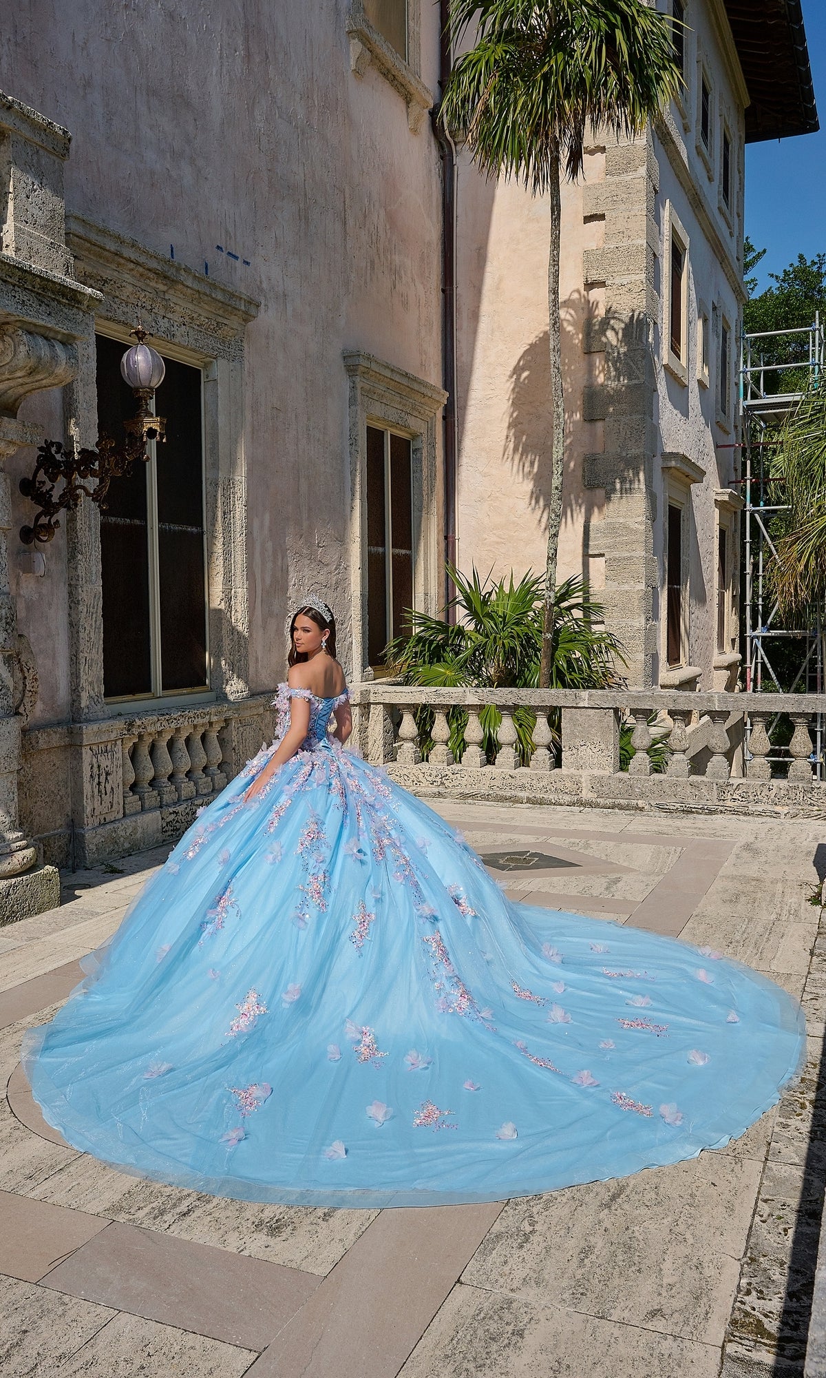 A young woman wears the Quinceanera Dress 54228 By Amarra, a light blue ball gown with 3D floral applique, as she stands on an outdoor stone terrace facing away from the camera with a historic building behind her.